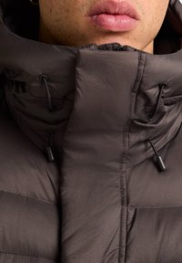 Giacca a vento marrone con colletto alto, dotata di lacci regolabili e texture liscia. Presenta una cerniera e pannelli cuciti.