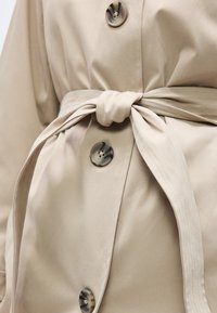 Κοντινό πλάνο μπεζ καμπαρντίνας με δεμένη υφασμάτινη ζώνη και μεγάλα κουμπιά τετραγωνικού σχήματος με μοτίβο χελώνας κατά μήκος του μπροστινού μέρους.