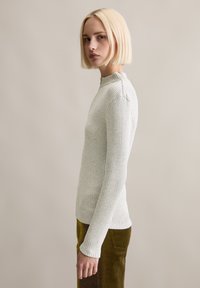 Blonde Frau mit geradem Bob-Haarschnitt, die einen hellgrauen, gerippten Pullover mit langen Ärmeln und olivgrünen Cordhosen trägt, steht seitlich.