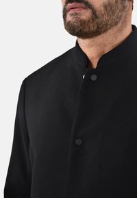 Giacca nera con colletto alla mandarino, realizzata in tessuto morbido. Presenta un design semplice con due bottoni neri visibili e una superficie liscia.