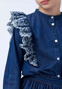 Denimová šaty s knoflíkovou přední stranou a volánkovými rameny s bílým krajkovým ozdobením. Tmavě modrá látka, řasený pas a dlouhé rukávy.