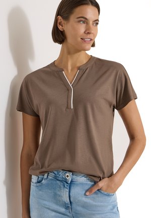 Femme aux cheveux bruns courts portant un haut marron à manches courtes avec un liseré blanc et un jean bleu clair, une main dans la poche.