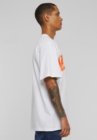 White cotton t-shirt with a loose fit, featuring an orange graphic design on the front. Short sleeves and a round neckline.