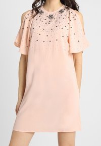 Světle broskvové šaty s odhalenými rameny, krátkými roztaženými rukávy a černými korálkovými ozdobami kolem výstřihu a horní části hrudníku.