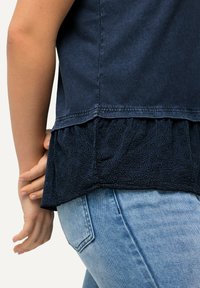 Parte superior de un oscuro color azul marino con un dobladillo bordado y con volantes; combinada con jeans de color azul claro. Presenta una tela texturizada y un sutil patrón floral en el dobladillo.
