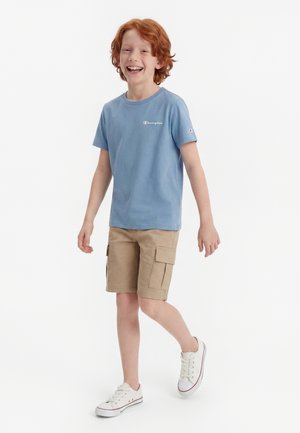 Smilende rødhåret dreng, der går, iført en blå Champion t-shirt, beige cargoshorts og hvide sneakers mod en hvid baggrund.