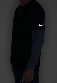 Person, der ein schwarzes Langarmshirt von Nike trägt, mit einem reflektierenden weißen Nike-Swoosh-Logo auf dem Oberarm, steht vor einem dunklen Hintergrund.
