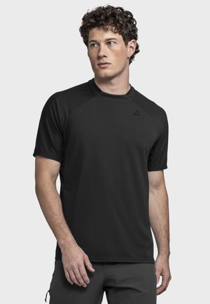 Junger Mann mit lockigem Haar, der ein einfaches schwarzes, kurzärmeliges Sportshirt und dunkle Hosen trägt, blickt seitlich vor einem weißen Hintergrund.