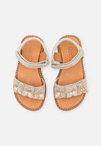 Sandales beiges avec des embellissements floraux sur la bride, une bride à la cheville réglable et une semelle intérieure beige. Matériau supérieur texturé.
