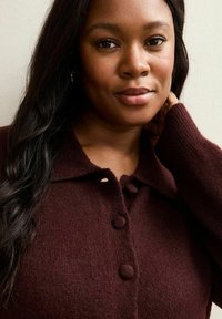 Burgundowa dzianinowa kardigan z kołnierzem, z trzema dużymi guzikami i teksturalnym wykończeniem. Model delikatnie pozuje z jedną ręką na szyi.