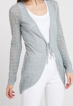 Jasnoszary otwarty sweter na drutach z wiązaniem z przodu, noszony na białej bluzce i zestawiony z białymi spodniami.