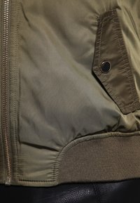Béžová bombera olivové barvy s hladkým nylonovým povrchem, předním zapínáním na zip, žebrovanými manžetami a zakulacenou kapsou s kovovým knoflíkem.