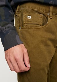Khaaki spodnie wykonane z denimu o gładkiej fakturze, z boczną kieszenią, srebrnymi detalami oraz dopasowanym krojem.