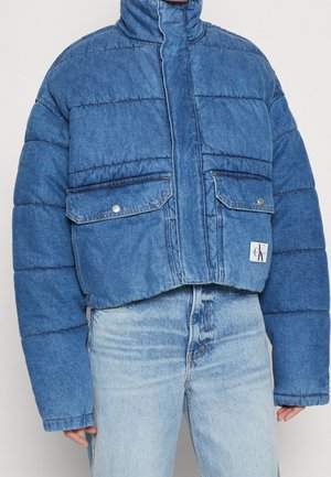 Niebieska, krótkia kurtka puchowa z denimu z wysokim kołnierzem, kieszeniami na zatrzaski z patkami oraz naszywką z logo Calvin Klein na dolnej przedniej części, noszona z jasnymi jeansami.
