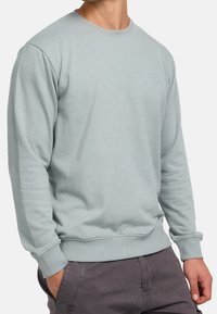 Hombre con sudadera de cuello redondo gris claro con puños y dobladillo acanalados, combinada con pantalones gris oscuro, de pie con una mano en el bolsillo.