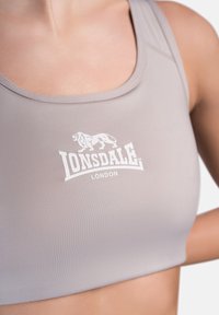Šviesiai pilkas sportinis liemenukas su lygiu paviršiumi, priekyje turintis spausdintą baltą Lonsdale logotipą ir apvalaus iškirpimo dizainą.
