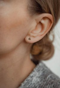 Goldene Ohrstecker in dreieckiger Form mit drei kleinen schwarzen Akzenten, am Ohrläppchen getragen, vor einem verschwommenen Hintergrund.