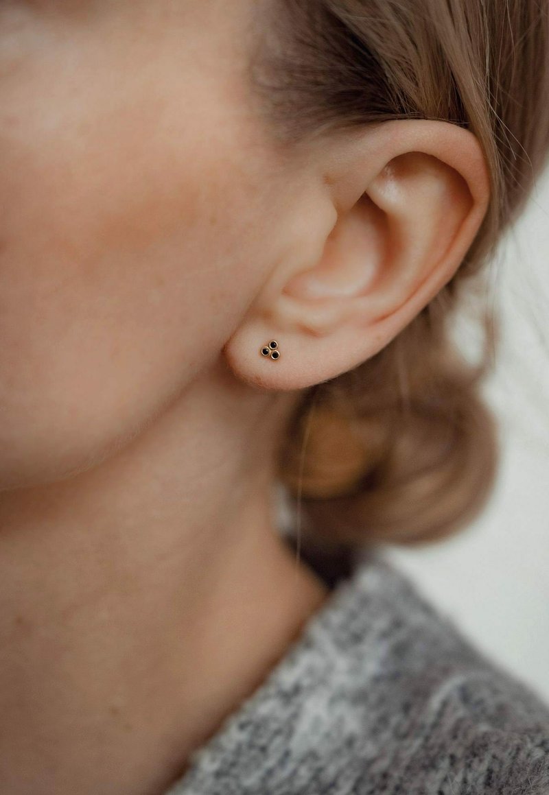 Goldene Ohrstecker in dreieckiger Form mit drei kleinen schwarzen Akzenten, am Ohrläppchen getragen, vor einem verschwommenen Hintergrund.