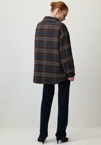 Srajčna jakna v karo vzorcu v odtenkih modre in rjave, z udobnim ohlapnim krojem, velikim ovratnikom in dolgimi rokavi. Kombinirana s temnimi hlačami.