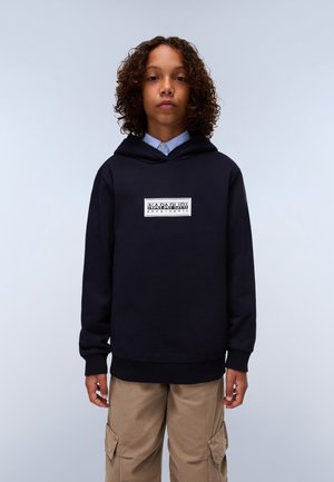 Sudadera navy con un bolsillo frontal y puños acanalados. Presenta un parche de logo blanco rectangular en el centro. Combinada con pantalones cargo beige.