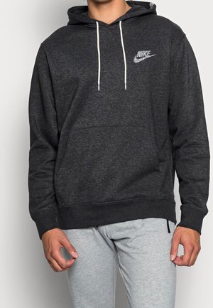 Mand iført en mørkegrå Nike-hoodie med hvide snore og lysegrå joggingbukser, stående mod en ensfarvet baggrund.