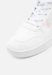Baskets blanches avec empeigne synthétique lisse, bout arrondi, semelle en caoutchouc texturée et accent irisé brillant sur le côté.