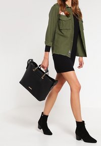 Svart tote-väska med guld detaljer, rektangulär form och dubbla handtag; slät yta, som kontrasterar med en olivgrön jacka och svart klänning.