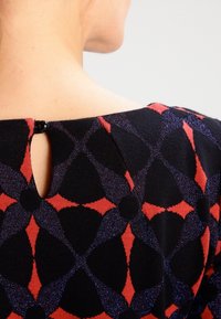 Kvinnas övre rygg iklädd en svart klänning med röd och lila geometrisk blommönster och en liten svart knapp vid halsringningen.