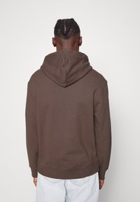 Sudadera de algodón marrón con un corte relajado, capucha con cordón y puños acanalados. Presenta una textura suave y lisa, con detalles de costura mínimos.