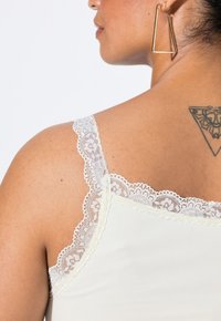 Top sin mangas color crema con delicado encaje blanco en los tirantes; tela suave y un tatuaje visible en el hombro de la espalda.