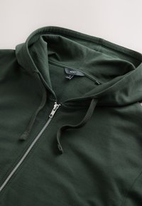 Donkergroene sweatvest met ritssluiting in zilver, een trekkoordkap en zachte katoenstof. De binnenkant heeft een eenvoudige, onopgesmukte uitstraling.