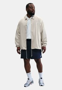 Veste légère beige, chemise blanche à col, shorts navy, et chaussettes blanches avec des chaussures de sport grises et rouges, se tenant contre un fond uni.