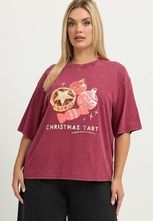 Femme blonde portant un t-shirt bordeaux ample avec un motif tartan de Noël et ornement, et un pantalon noir sur un fond uni.