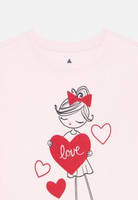Light pink t-shirt featuring a cartoon girl with a red bow, holding a red heart with "love" text, surrounded by smaller red hearts.