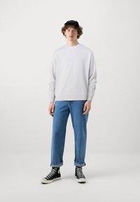 Felpa grigia chiaro con maniche lunghe abbinata a jeans a gamba dritta blu e sneakers alte nere, in piedi davanti a uno sfondo bianco.