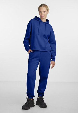 Blauw katoenen hoodie met trekkoorden en een ontspannen fit, gecombineerd met bijpassende blauwe joggingbroeken. Zwarte sneakers maken de outfit compleet.