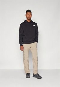 Camisola preta com capuz, calças cargo castanhas e sapatos de caminhada pretos. Logótipo notável na camisola e bolsos nas calças.