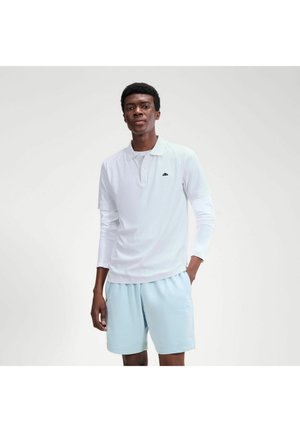 Junger Mann, der ein weißes Langarm-Poloshirt und hellblaue Shorts trägt, steht mit einer Hand in der Tasche vor einem einfarbigen Hintergrund.