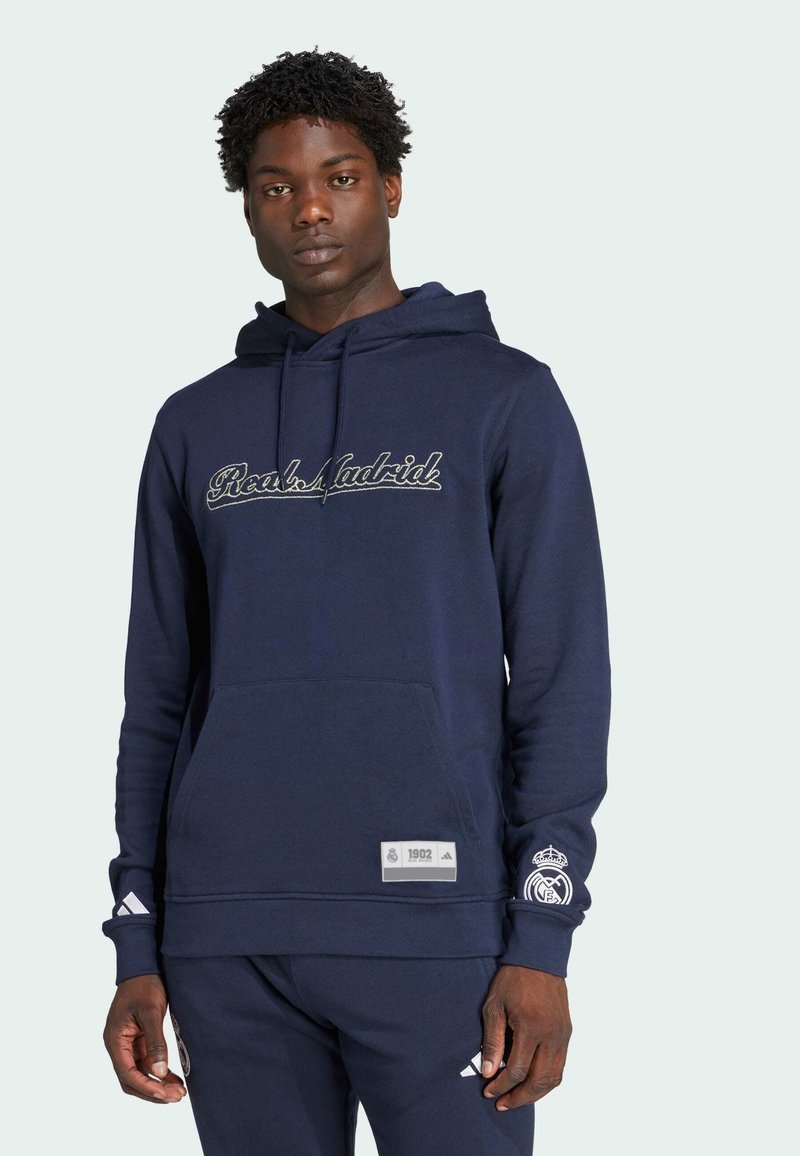 Mörkblå sweatshirt i mjukt material, med en ficka framtill, broderad text "Real Madrid" och logotypdetaljer på ärmarna.