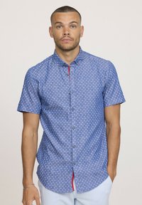 Chemise à manches courtes en tissu bleu clair avec de petits motifs blancs, une subtile touche de rouge sur la patte de boutonnage et des boutons en métal.