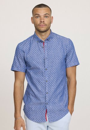 Chemise à manches courtes en tissu bleu clair avec de petits motifs blancs, une subtile touche de rouge sur la patte de boutonnage et des boutons en métal.