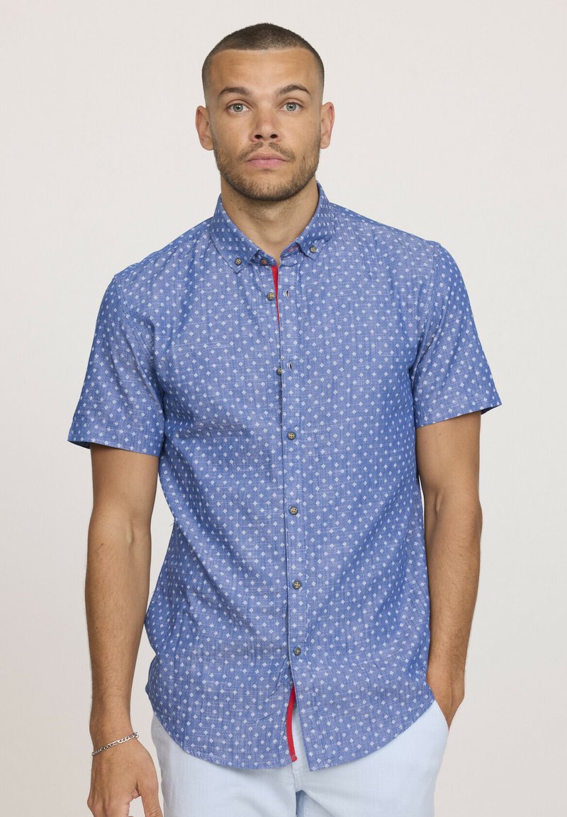 Chemise à manches courtes en tissu bleu clair avec de petits motifs blancs, une subtile touche de rouge sur la patte de boutonnage et des boutons en métal.