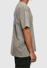 Mand iført en løs grå acid-wash t-shirt med korte ærmer, stående sidelæns, parret med sorte bukser mod en ensfarvet baggrund.