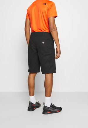 Homme portant une chemise à manches courtes orange vif, un short noir et des baskets noires avec des chaussettes blanches, debout contre un fond clair uni.