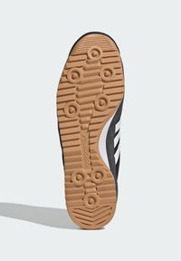 Suela negra de zapatilla con un patrón de goma de goma y ranuras centrales. Presenta tres rayas blancas en el costado y un material texturizado en la parte superior.