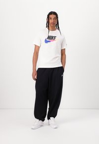 Balta medvilninė marškinė su spalvingu Nike logotipu, derinama su laisvo kirpimo juodomis treningų kelnėmis ir baltais sportiniais bateliais. Paprasta, laisva apranga.