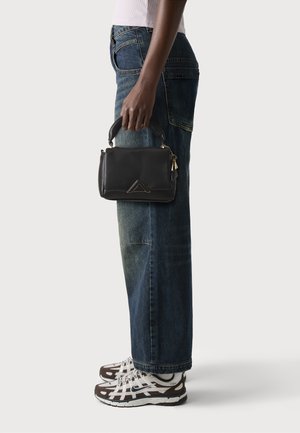 Black leather handbag with a structured design, top handle, gold hardware, and a triangular accent, held with dark denim wide-leg jeans and sneakers.