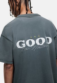 Grijze oversized t-shirt met korte mouwen, voorzien van "GOOD For Nothing" in het wit met groene steraccenten op de achterkant.