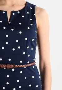 Marineblaues Polka-Dot-Kleid mit leichtem V-Ausschnitt, aus Baumwolle gefertigt und mit einem braunen geflochtenen Gürtel in der Taille akzentuiert.