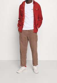 Rode ribfluwelen overhemd over een wit T-shirt, gecombineerd met bruine ribfluwelen broek en lichte sneakers. Details van de gestructureerde stof zijn zichtbaar.